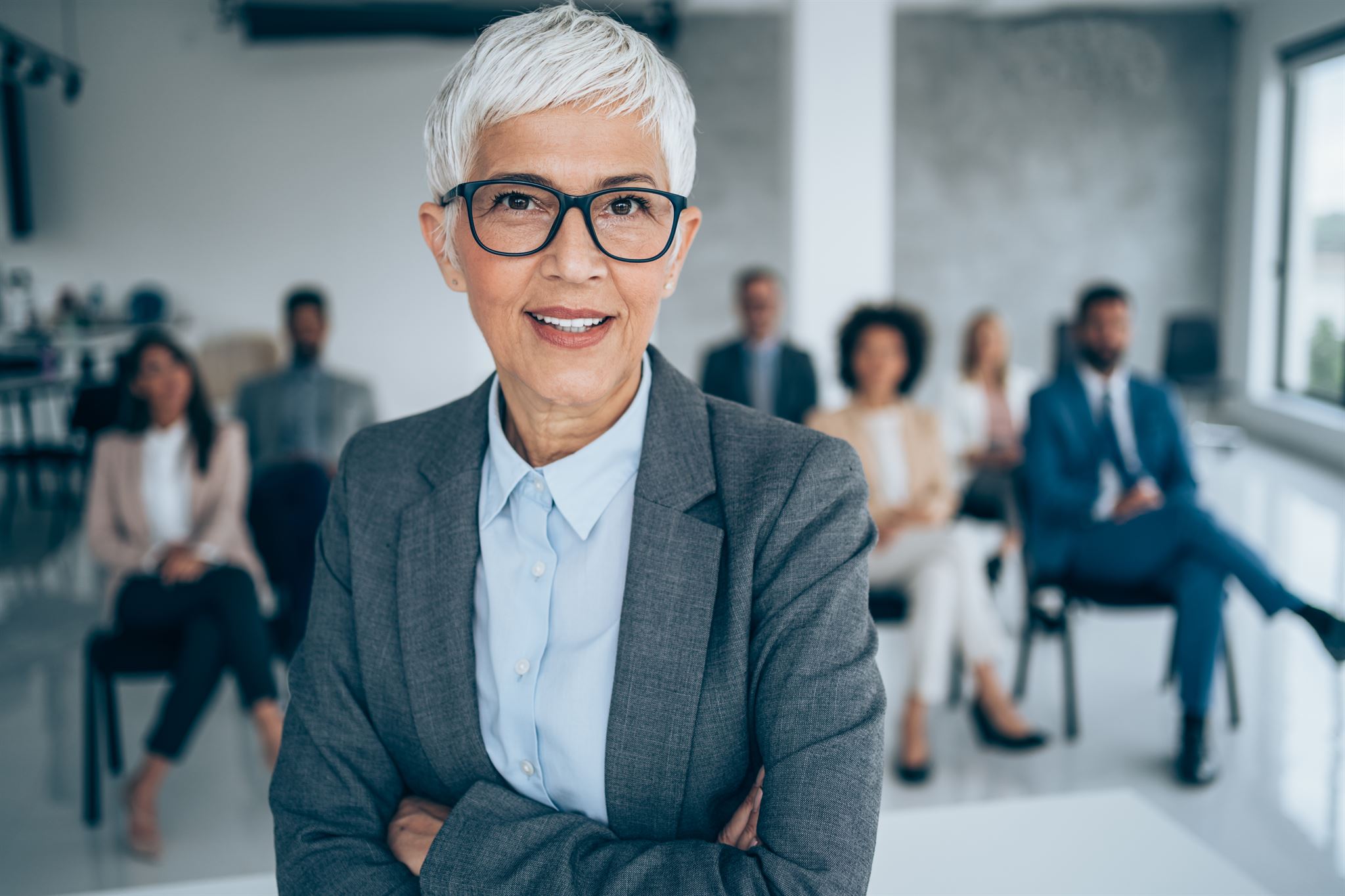 Short-haired-woman-in-glasses-and-suit-looking-at-camera-and-standing-in-front-of-seated-colleagues-in-conference-hall